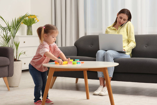 Woman working remotely at home. Mother talking on smartphone and using laptop while her daughter playing at desk - Zdjęcie, obraz