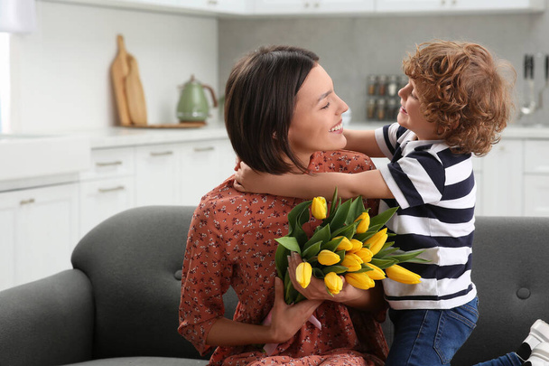 Little son congratulating his mom with Mother`s day at home. Woman holding bouquet of yellow tulips - 写真・画像