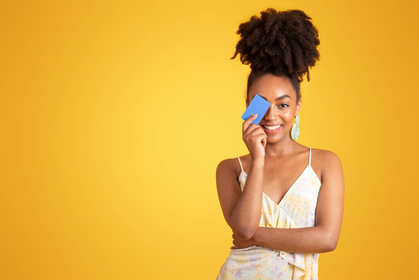 Счастливая молодая африканская американка в платье, закрывающая глаза кредиткой, изолированная на жёлтом фоне студии. Магазины и продажа, финансы, деньги, образ жизни, реклама и предложение - Фото, изображение
