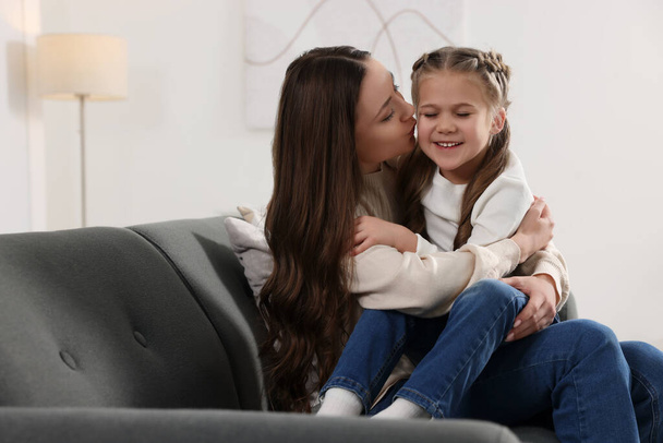 Happy mother kissing her cute daughter on sofa at home - Foto, Bild