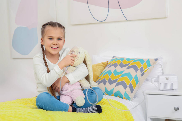 Little girl with toy bunny and nebulizer for inhalation on bed at home - Foto, afbeelding