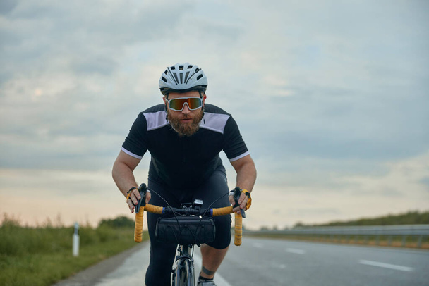 Dynamic image of young bearded man , cyclist in helmet riding bike on empty road in the evening. Concentration. Concept of sport, hobby, leisure activity, training, health, speed, endurance, ad - Foto, Bild