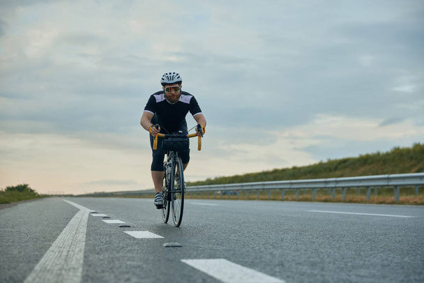 Dynamic image of young, sportive, bearded man, cyclist in helmet riding bicycle on empty road in the evening. Concept of sport, hobby, leisure activity, training, health, speed, endurance, ad - Photo, Image