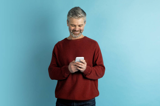 Cheerful handsome grey-haired bearded middle aged man in red sweater using cell phone and smiling, isolated on blue background, copy space. Chatbot, online assistant, AI in daily life - Photo, Image