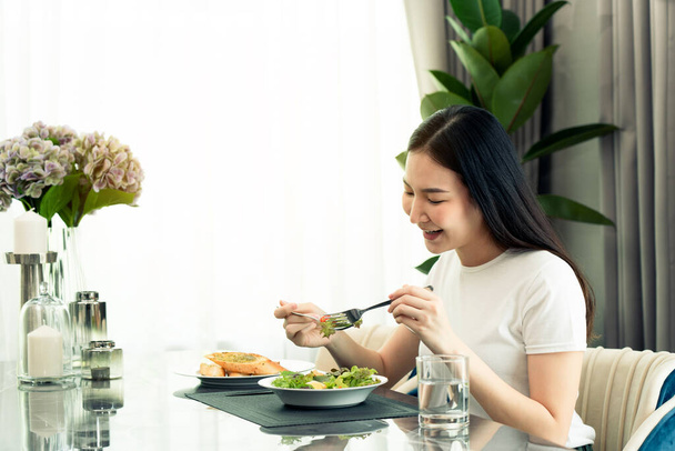 Asian young woman smiling as she scoops a salad on a plate and eats happily at home. - Фото, изображение