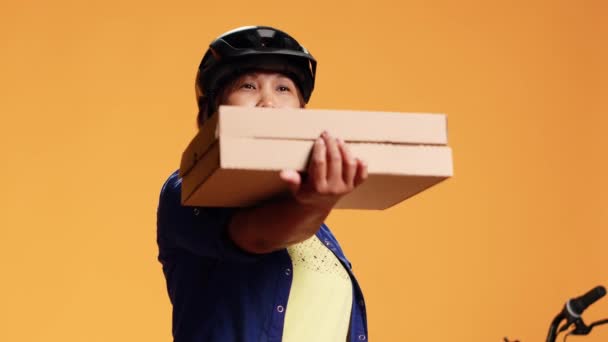 Hip level close up shot of cheerful courier looking at camera, showing approval gesture while delivering takeout meals. Friendly cyclist offering pizza to customer, isolated over studio background - Footage, Video