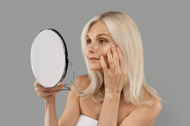 Skin Aging. Attractive Mature Woman Holding Mirror And Looking At Her Wrinkles Near Eyes, Beautiful Senior Female Checking Face, Noticing Age Changes, Standing Wrapped In Towel On Grey Background - Zdjęcie, obraz