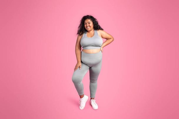 Ganztägige Aufnahme einer glücklichen schwarzen übergewichtigen Dame in Sportbekleidung, die isoliert auf rosa Hintergrund in die Kamera lächelt. Pilates und Körperpflege, Sport und Angebot, Fitness und Workout - Foto, Bild