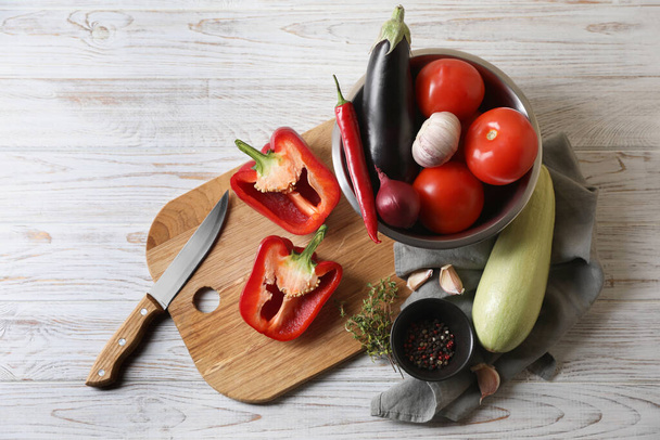Cooking delicious ratatouille. Fresh ripe vegetables, knife and bowl on white wooden table, flat lay - Fotó, kép