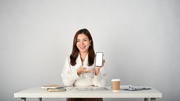 A beautiful and smiling young Asian businesswoman sits at her desk and shows her smartphone white screen mockup to the camera. isolated white studio background - Valokuva, kuva