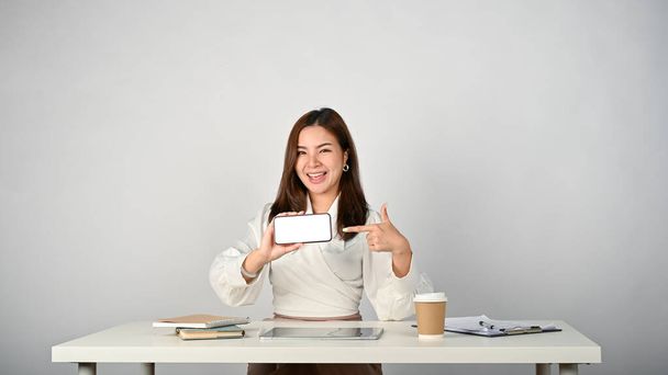 Wesoła młoda azjatycka bizneswoman siedzi przy biurku, wskazując palcem na ekran smartfona. - Zdjęcie, obraz