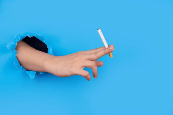 Hand through paper holding cigarette on blue background - Foto, afbeelding