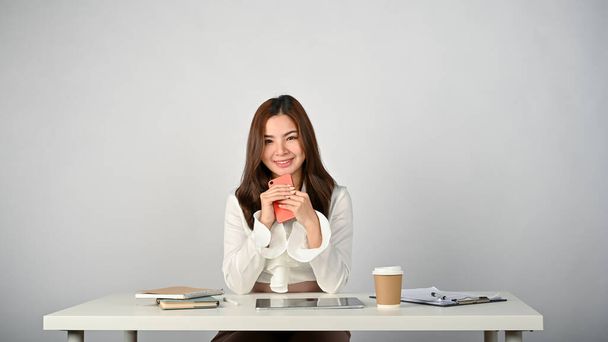 Egy gyönyörű és elbűvölő fiatal ázsiai üzletasszony vagy női irodai dolgozó ül az asztalánál okostelefonjával a kezében. izolált fehér háttér - Fotó, kép