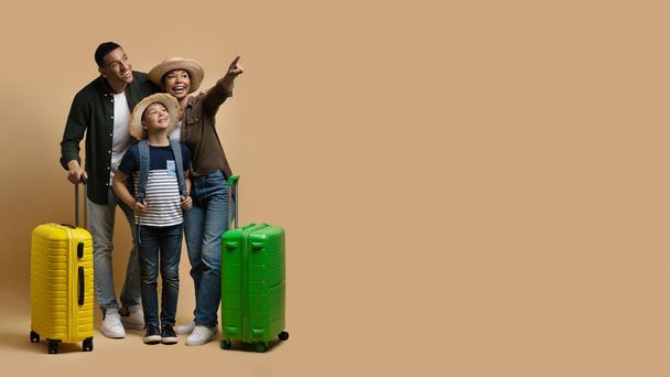 Családi nyaralás, nyaralás együtt koncepció. Vidám fekete családi turisták utaznak együtt, boldog apa, anya és iskolás korú fiú bőröndökkel rámutatva a fénymásolótérre bézsen, panorámán - Fotó, kép