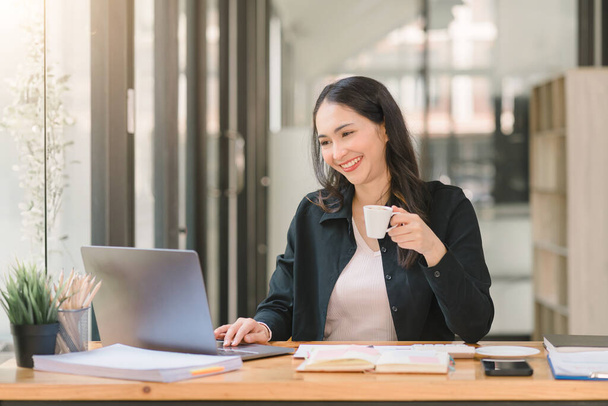 An attractive, young, and confident mixed-race asian businesswoman is sitting at the office table, enjoying an espresso shot in a modern office - Photo, Image
