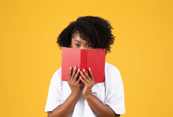Frohe schwarze Millennial-Dame in lässigem Look von hinter Buch, lesen isoliert auf orangefarbenem Studiohintergrund. Lernen, Wissen und Bildung, Hausaufgaben, Entspannung und Hobbys, Spaß - Foto, Bild