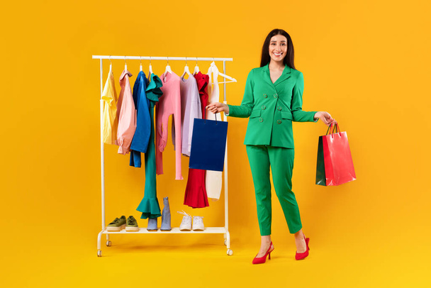Happy young stylish european woman posing with shopper bags near clothing rail, satisfied with great sales, yellow studio background, full length shot, copy space - Photo, Image