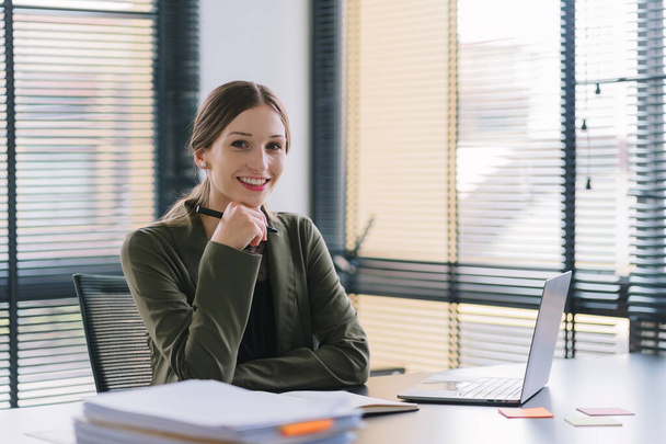 A portrait of a millennial businesswoman in a stylish dress is sitting at desk in modern office, using a laptop computer. She appears to be a successful finance manager who is planning work projects - 写真・画像