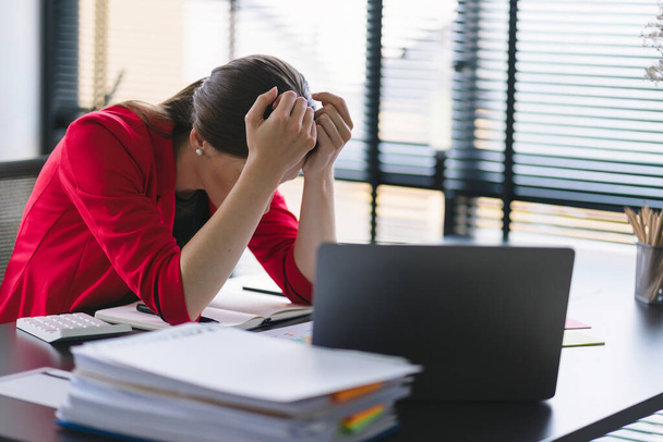 La estresada y agotada empresaria milenaria es vista sentada en el escritorio de su oficina con la mano en la cabeza, indicando un día de trabajo duro donde está sobrecargada de trabajo - Foto, imagen