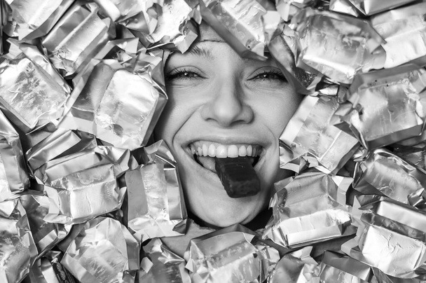 The face of a Caucasian woman surrounded by candy wrappers. The girl eats a bar of chocolate. Monochrome - Zdjęcie, obraz