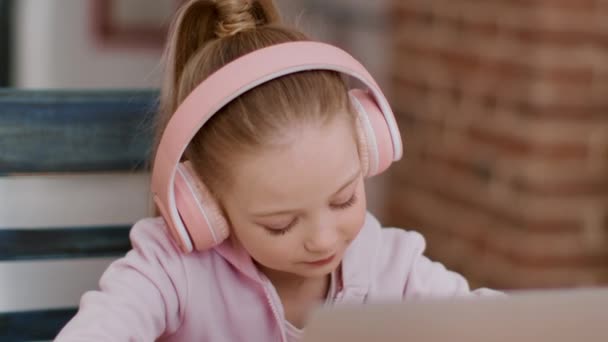 Grundlegende Online-Bildungskonzept. Nahaufnahme Porträt eines intelligenten kleinen Mädchens mit Kopfhörern, das zu Hause lernt, Unterricht am Laptop verfolgt und Aufgaben schreibt, freier Raum - Filmmaterial, Video