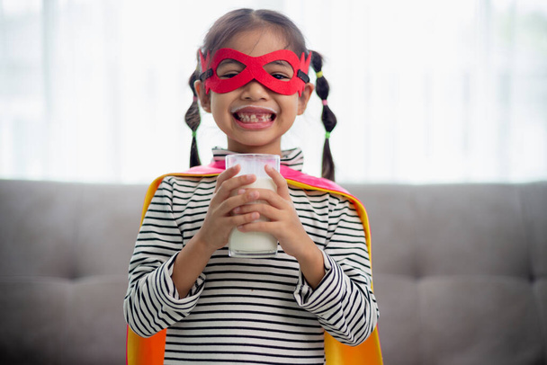 Азиатская маленькая девочка в костюме супергероя пьет молоко дома. Здоровое питание. - Фото, изображение