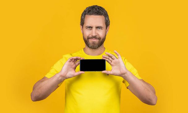 cheerful man showing smartphone app isolated on yellow. man showing smartphone app on background. man showing smartphone app in studio. photo of man showing smartphone app. - Photo, Image