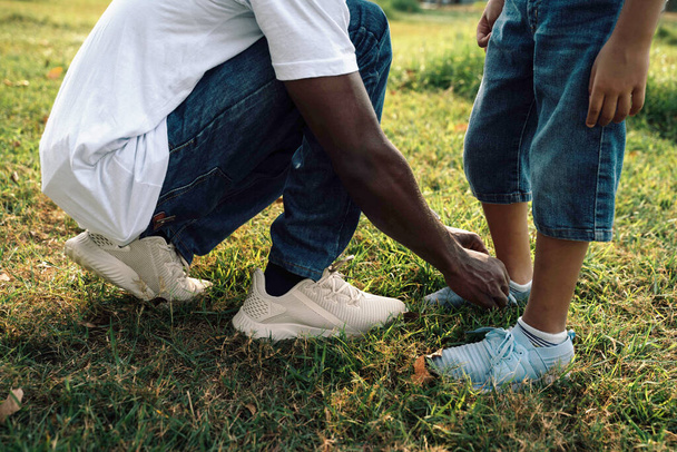 Warmer Sonnentag im Park: Afroamerikanischer Vater hilft Sohn beim Schnüren von Schuhen - Foto, Bild