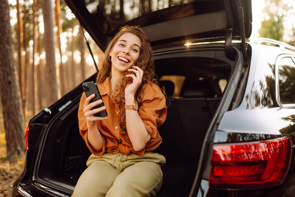 Красива жінка сидить у багажнику машини в лісі і використовує смартфон. Поняття подорожі автомобілем, активний спосіб життя. - Фото, зображення
