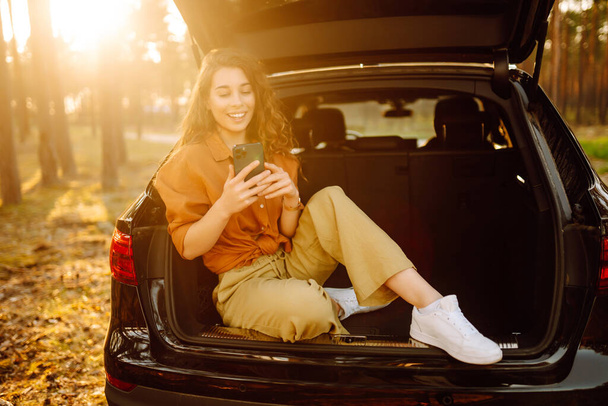 Beautiful woman sits in the trunk of a car in the forest and uses a smartphone. The concept of traveling by car, active lifestyle. - 写真・画像