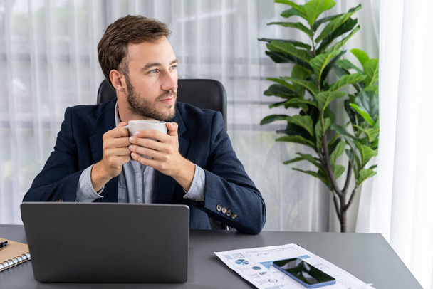 Zakenman werkt aan zijn laptop en drinkt koffie aan zijn bureau in de werkruimte. Smart executive nemen koffiepauze na het onderzoeken van financiële gegevens en marketing plan op zijn zakelijke laptop. Entiteit - Foto, afbeelding