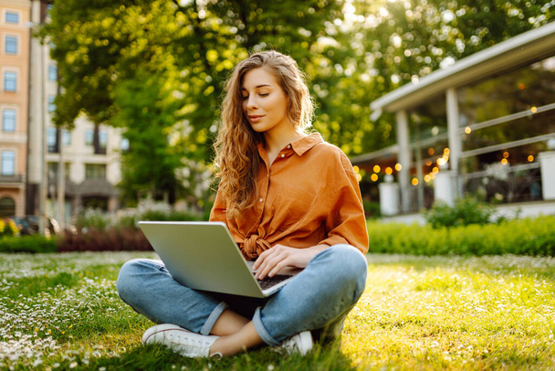 Beautiful female freelancer sits on a green meadow with a laptop. Online education. Happy woman running outdoors enjoying the sun. Freelancing concept. - Фото, изображение
