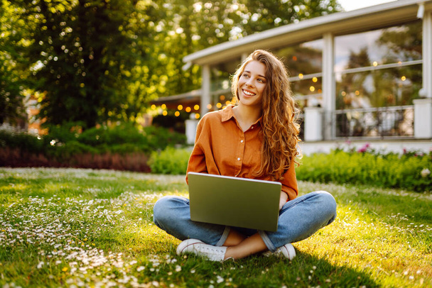 Beautiful female freelancer sits on a green meadow with a laptop. Online education. Happy woman running outdoors enjoying the sun. Freelancing concept. - Foto, imagen