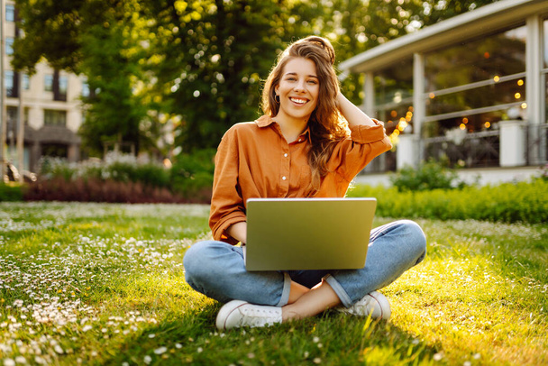 Beautiful female freelancer sits on a green meadow with a laptop. Online education. Happy woman running outdoors enjoying the sun. Freelancing concept. - Foto, Imagem