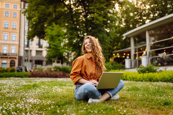 Hermosa freelancer femenina se sienta en un prado verde con un ordenador portátil. Educación en línea. Mujer feliz corriendo al aire libre disfrutando del sol. Concepto de autónomo. - Foto, imagen