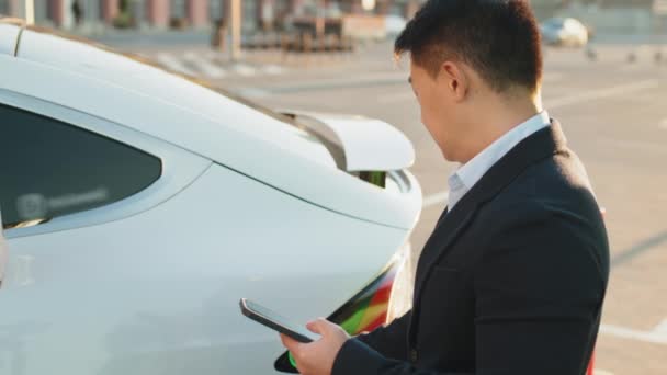 Siyah takım elbiseli Asyalı bir adamın beyaz lüks arabayı şarj edip modern akıllı telefon kullanmasına yakın çekim. Elektrikli araç teknolojik şarj konsepti. Arabaları şarj eden ve akıllı telefonları kullanan adama yaklaş.. - Video, Çekim