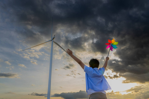 Progressive young asian boy playing with wind pinwheel toy in the wind turbine farm, green field over the hill. Green energy from renewable electric wind generator. Windmill in the countryside concept - Photo, image