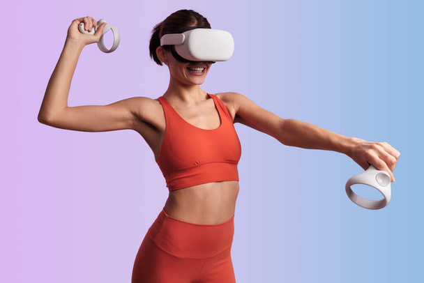 Cheerful young female athlete with dark hair in sportswear and VR goggles playing virtual tennis with gamepads and smiling against two color background - Foto, Imagem