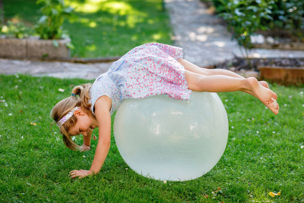 Pieni söpö tyttö tekee harjoituksia pallo ulkona. Aktiivinen esikoululainen pitää hauskaa urheilun kanssa. Ulkona lasten toimintaa kesällä - Valokuva, kuva