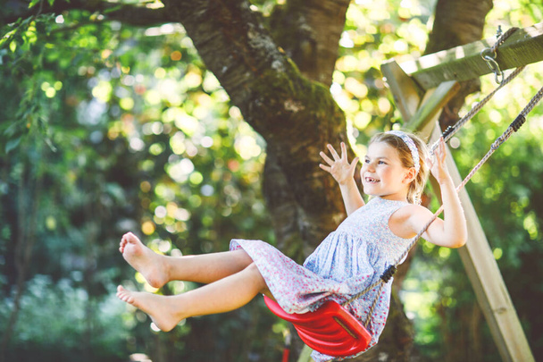 Menina pré-escolar pequena feliz se divertindo no balanço no jardim doméstico. Criança saudável bonito balançando sob árvores florescendo no dia ensolarado da primavera. Criança rindo e chorando - Foto, Imagem