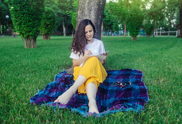 Καλλιτεχνική θεραπεία. Μια γυναίκα ζωγραφίζει στο πάρκο. Το κορίτσι κάθεται με την πλάτη πάνω σε ένα δέντρο και κοιτάζει σε απόσταση. Αποκατάσταση του νευρικού συστήματος με τη βοήθεια της σχεδίασης. Έννοια της χαλάρωσης και της διαλογιστικής κατάστασης - Φωτογραφία, εικόνα