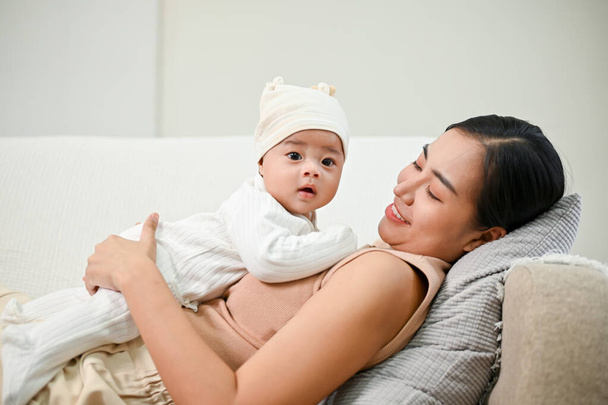 Hermosa madre asiática sosteniendo a su bebé mientras se relaja en el sofá, pasando tiempo con su hijo.  - Foto, Imagen