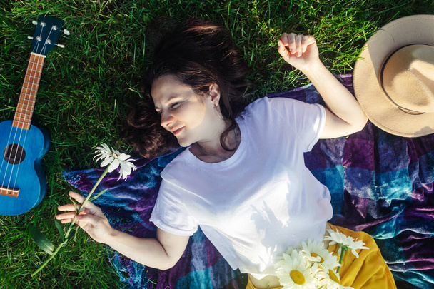 女性は紫の毛布にかかっている。彼女の体にはデイジーの花束です。公園内の瞑想状態。ウクレレを背景に、小さなギター。神経系治療の概念,自然療法 - 写真・画像