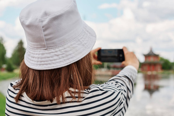 Γυναίκα στην καλοκαιρινή λίμνη, λίμνη ποταμού, φωτογράφηση τοπίου στο smartphone, θέα από πίσω, καταγραφή βίντεο στο Διαδίκτυο, γυναίκα σε ριγέ πουκάμισο και Παναμά. - Φωτογραφία, εικόνα