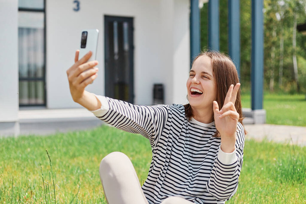 Brown haired woman making selfie photo on mobile phone, stylish happy smiling woman in striped shirt, posing on the background of green lawn and modern house, - Foto, Bild