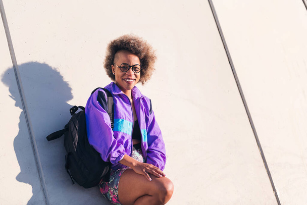 portrait of a smiling african american young woman in sports clothes leaning on an urban wall, concept of active lifestyle and urban sport, copy space for text - Φωτογραφία, εικόνα