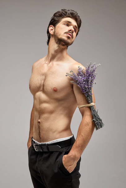 Мускулистый мужчина модель без рубашки с букет лаванды позирует на сером фоне. Концепция искусства, красоты, моды, праздников и рекламы. Мужественность. - Фото, изображение