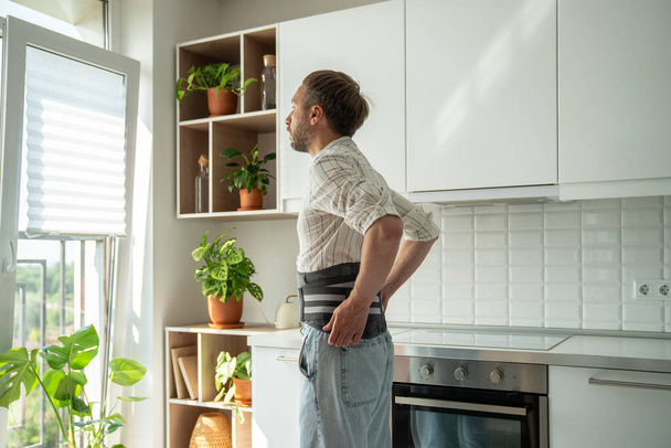 Beteg férfi otthon a konyhában, háttámaszt visel fűző a hát alsó részén a sérv kezelésének súlyosbodása során, műtét utáni felépülés. Hátfájás, gerinc egészségügyi problémák koncepció.  - Fotó, kép
