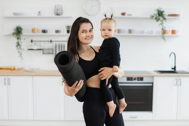 Portrait of joyful caucasian lady with yoga mat in hand carrying little baby girl while standing in spacious kitchen. Happy mother and adorable daughter in athletic wear planning home workout. - 写真・画像