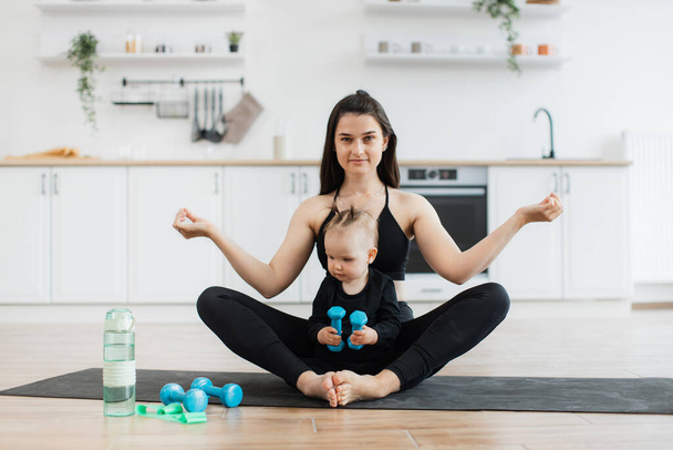 Full length view of relaxed female sitting in yoga pose with little child on rubber mat in modern apartment. Fit mother exercising with fingers in gyan mudra while cute daughter testing dumbbells. - 写真・画像